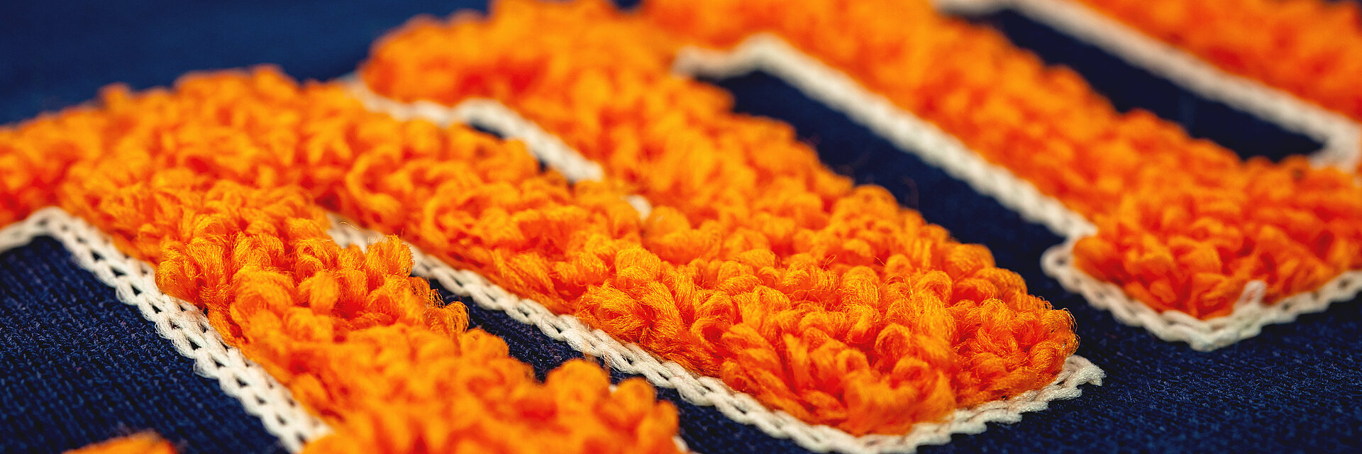Close-up of vibrant orange chenille embroidery on navy fabric, highlighting the soft volume and raised texture of the stitched lettering.